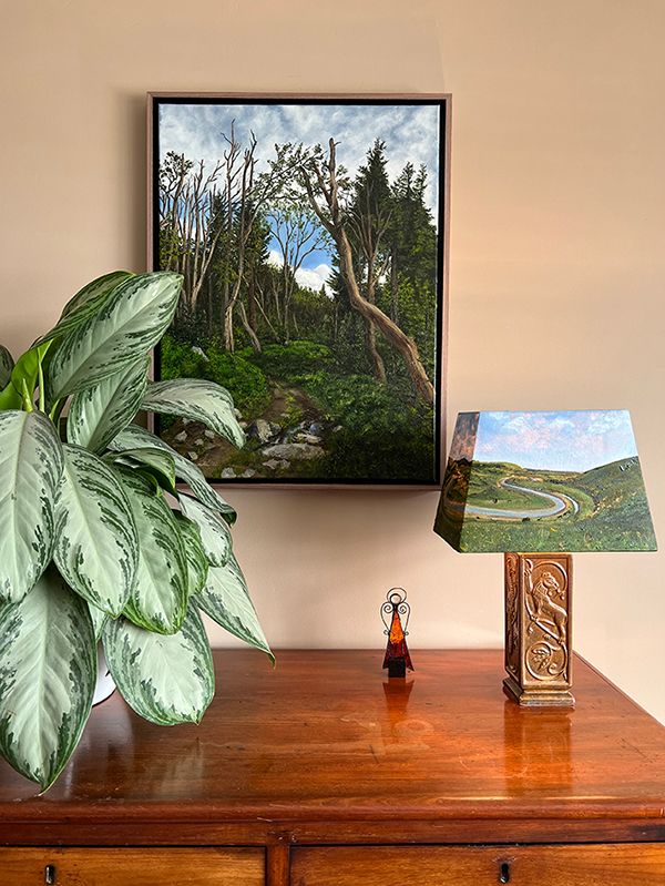 A desk with a painting of a landscape, a painted lampshade and plant on top of it. 